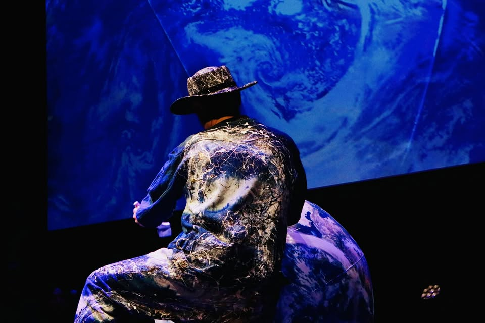 Shibuki Takakura in blue stage light, seen from behind in patterned clothing